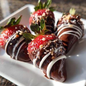 Four chocolate covered strawberries on a white plate, drizzled with white chocolate and sprinkled with nuts.