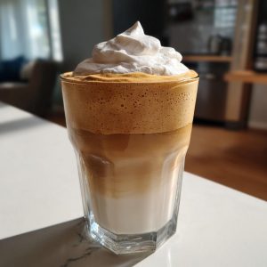 A glass of iced Dalgona Coffee topped with a generous swirl of whipped cream.
