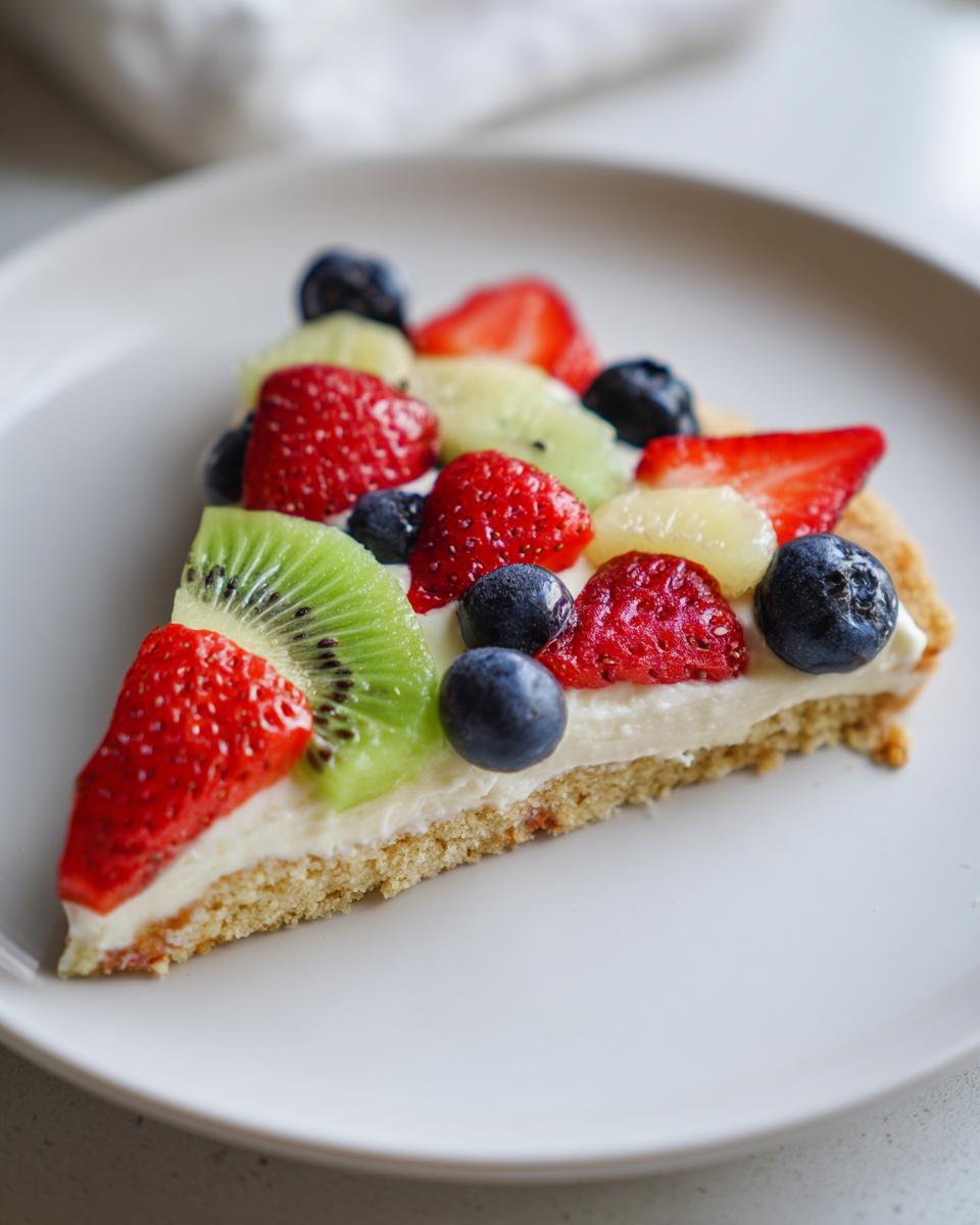 A slice of fruit pizza sugar cookie topped with strawberries, kiwi, and blueberries on a white plate.