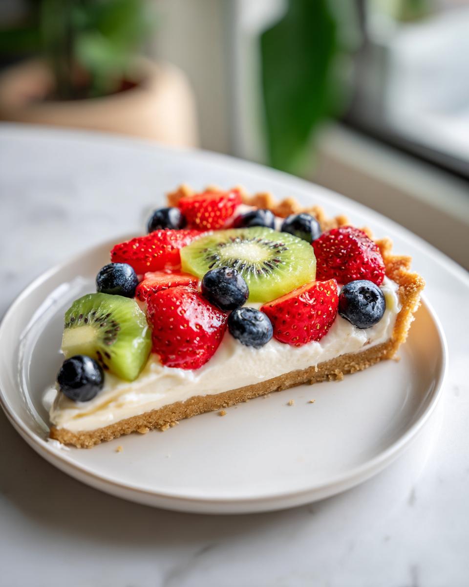 A slice of fruit pizza sugar cookie topped with strawberries, kiwi, and blueberries on a white plate.