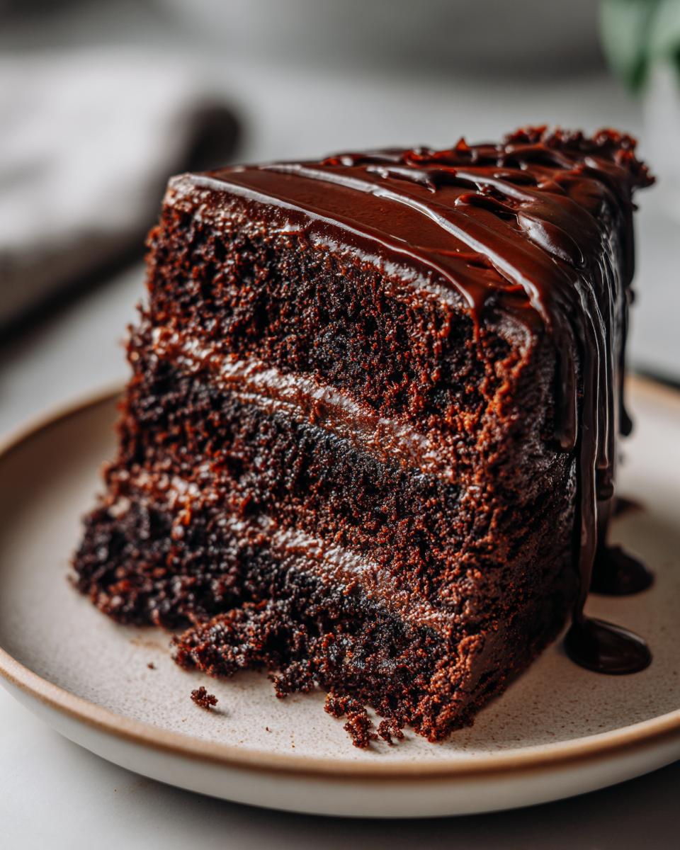 A decadent slice of Vegan Chocolate Cake, drizzled with chocolate ganache, sits on a plate.