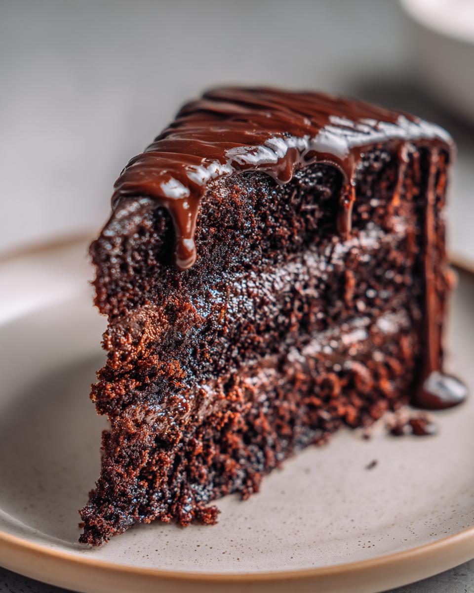 A decadent slice of Vegan Chocolate Cake topped with rich, dripping chocolate ganache on a plate.