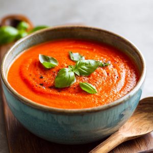 A close-up of a bowl of creamy tomato basil soup, garnished with fresh basil leaves and black pepper.