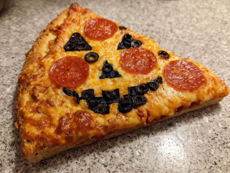 A slice of Jack-o'-lantern pizza decorated with pepperoni for eyes and mouth, and black olives for the nose and details.