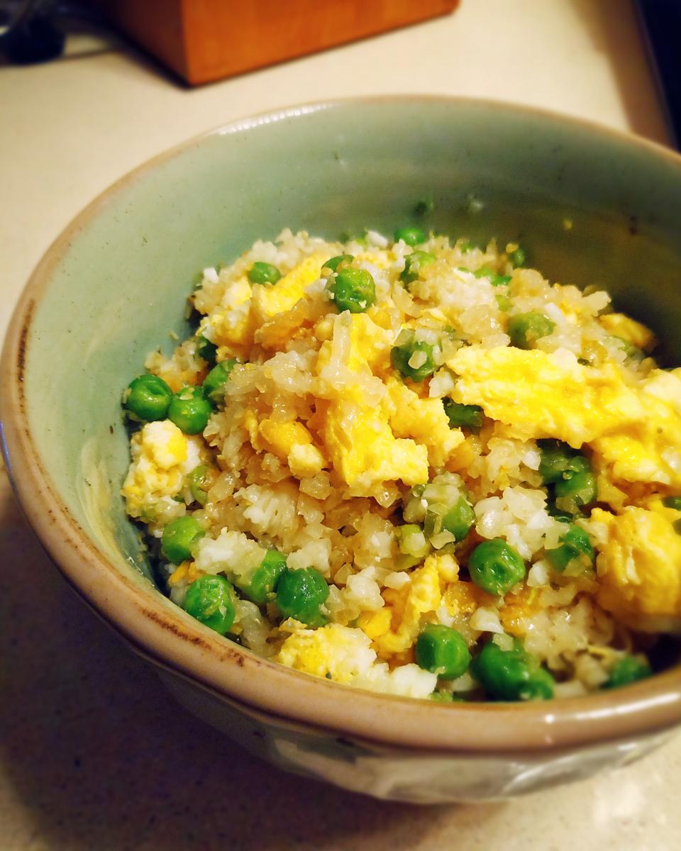 A close-up of a bowl of cauliflower fried rice mixed with bright green peas and scrambled egg.