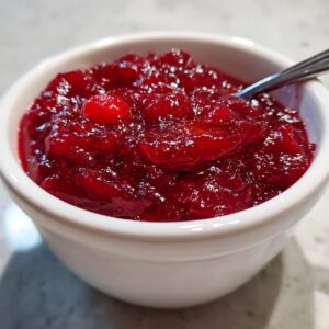 A close-up of vibrant, homemade cranberry sauce in a white bowl, with a spoon resting inside.
