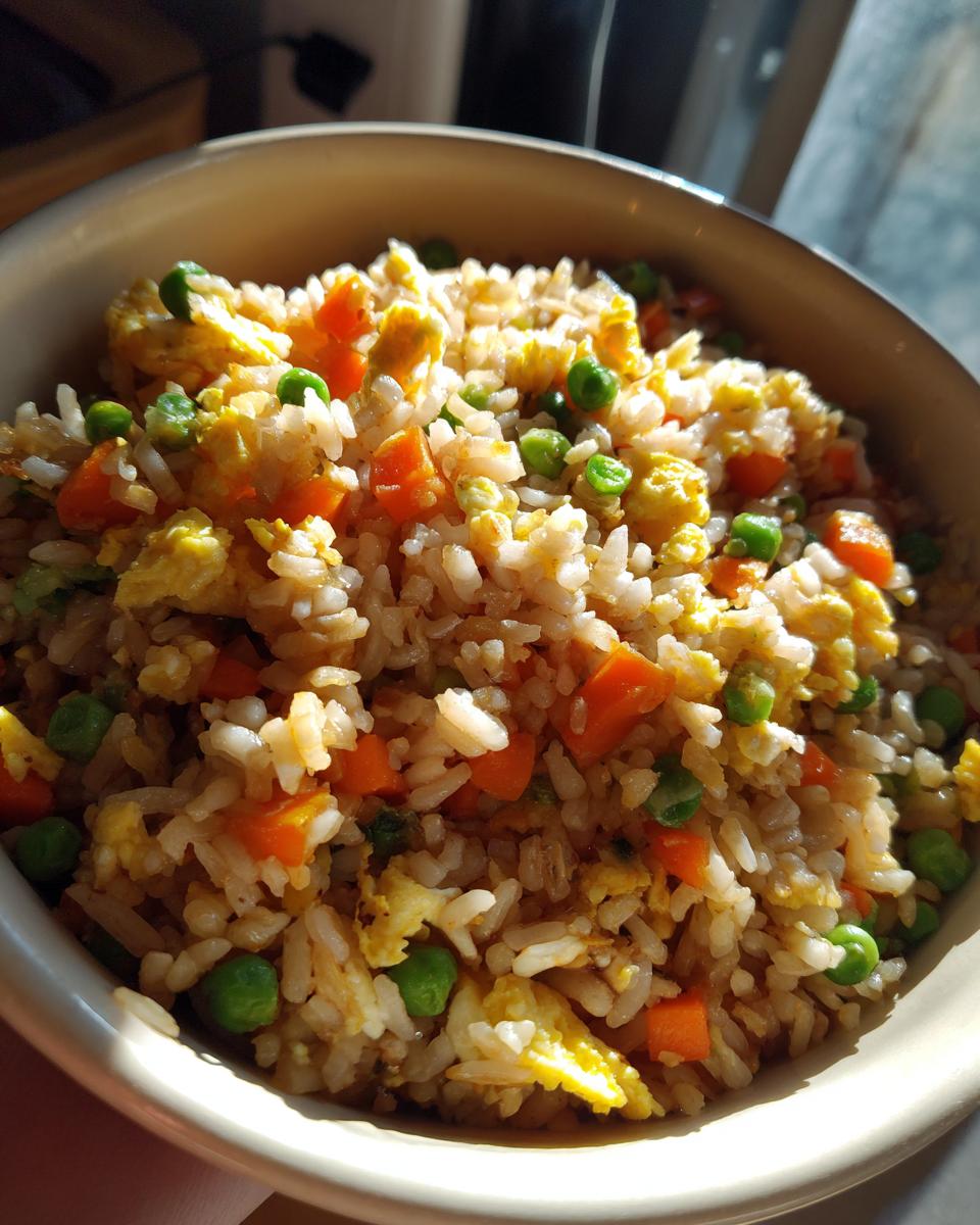 A close-up bowl of homemade fried rice, featuring rice, scrambled egg, peas, and diced carrots.