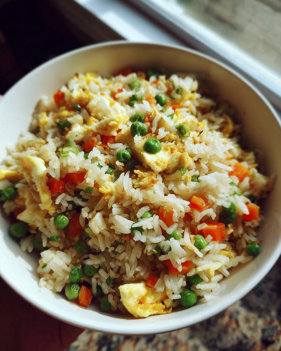 A close-up of a bowl filled with fluffy fried rice, studded with vibrant green peas, diced carrots, and scrambled egg pieces.