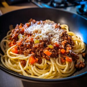 A close-up of a bowl of Spaghetti Bolognese topped with rich meat sauce, diced carrots, celery, and grated Parmesan cheese.