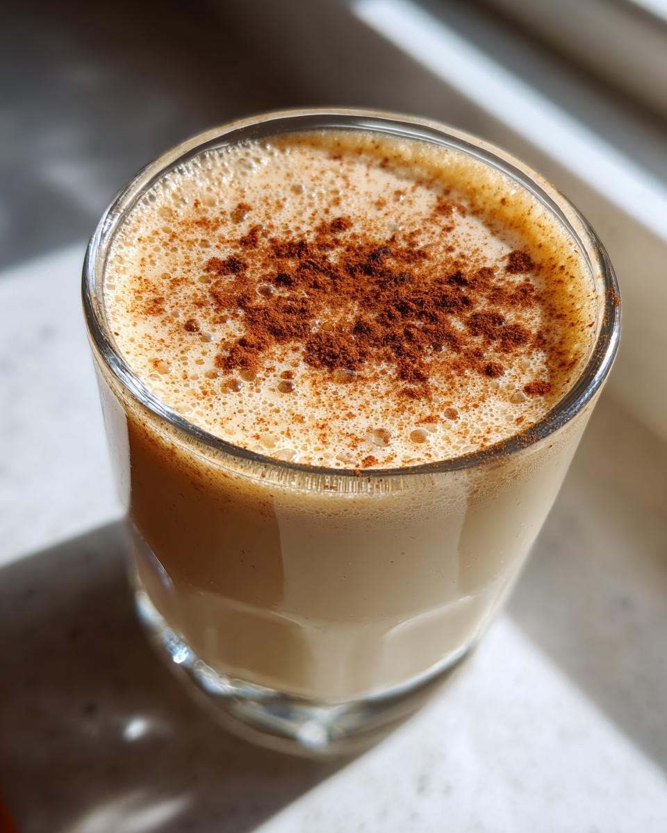 A close-up of a glass of creamy Coquito topped with a sprinkle of cinnamon.