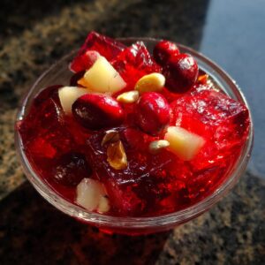 A close-up of a glass bowl filled with bright red Cranberry Jell-O salad, topped with whole cranberries, diced apples, and nuts.