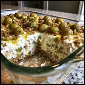 A serving spoon has taken a scoop out of the creamy dirty martini dip, revealing layers topped with whole green olives.