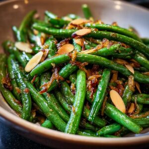 A close-up of vibrant green beans almondine, glistening and topped with toasted slivered almonds and shallots.