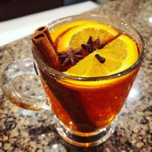 A clear glass mug filled with warm mulled cider, garnished with orange slices, cinnamon sticks, star anise, and cloves.