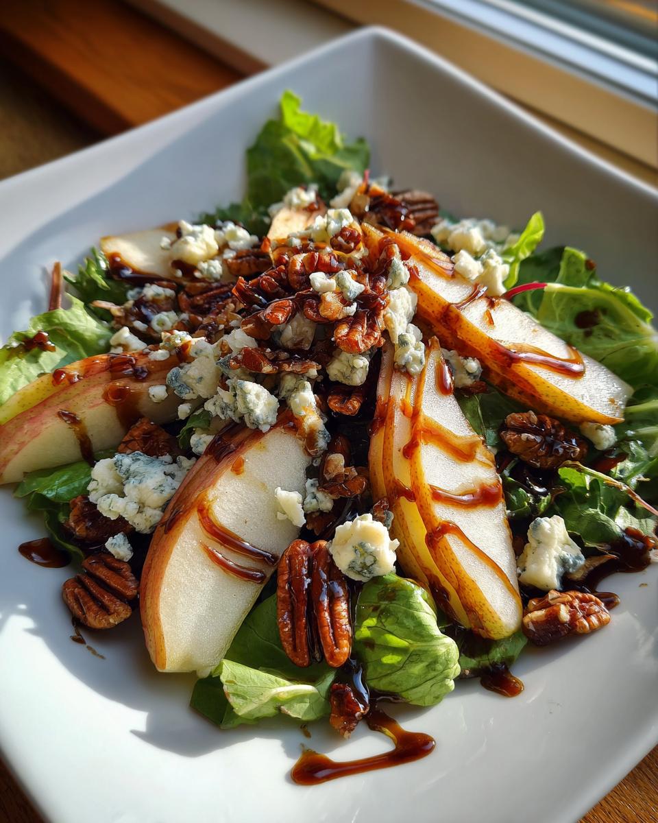 A refreshing pear salad featuring sliced pears, crumbled blue cheese, candied pecans, and a balsamic glaze on a bed of mixed greens.