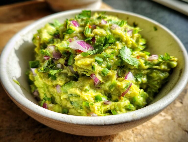 A close-up of a bowl filled with chunky, fresh spicy guacamole, garnished with red onion and cilantro.
